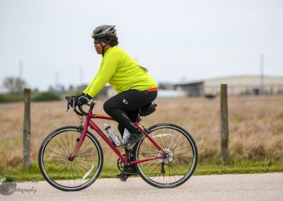 Bright yellow cyclist helmet, sunglasses, and high-visibility jacket riding a pink bike on a rural trail.