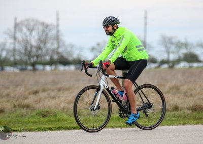 Man riding a bicycle on a paved trail during daytime, wearing a bright green jacket and safety gear.