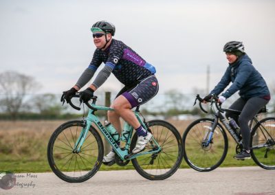 Bright blue Bluebonnet Express mountain bike with two cyclists riding outdoors on a cloudy day.