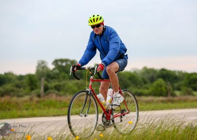 Older man biking on road in outdoor setting, wearing helmet and sunglasses, enjoying a ride with Bluebonnet Express bike.