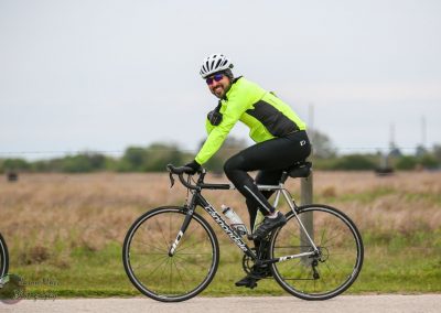 Vibrant cyclist riding a black Cannondale bike on a flat rural road, wearing neon green and black cycling gear.