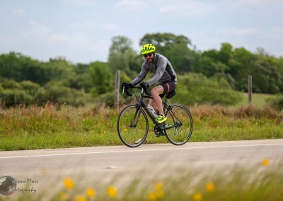 Vibrant cyclist riding a bike on a scenic rural road in spring, wearing a yellow helmet and black cycling gear, promoting Bluebonnet Express.
