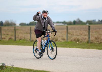 Bright man riding Bluebonnet Express bike, smiling, in rural open landscape with cloudy sky.