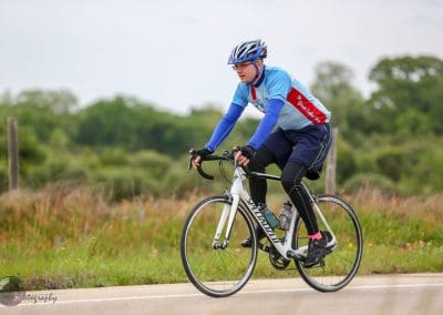 Vibrant cyclist in Bluebonnet Express jersey riding a Specialized road bike on a rural trail.