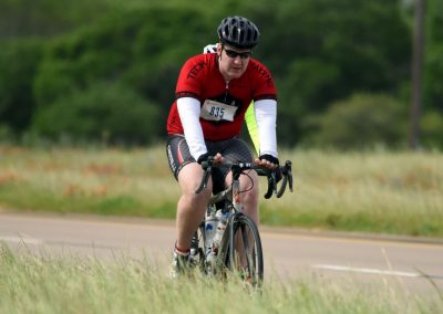 Cyclist riding on a rural road during a bike race, wearing a red jersey and helmet.