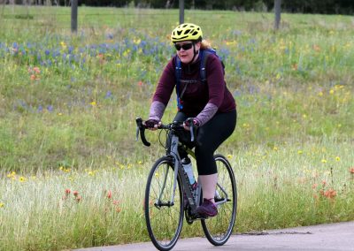 Bright woman riding a bicycle along a scenic trail with wildflowers, promoting Bluebonnet Express cycling tours and outdoor adventure.