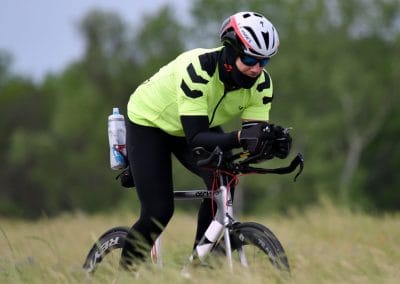High-performance cyclist riding on a grassy trail, wearing a helmet, sunglasses, and bright cycling gear.