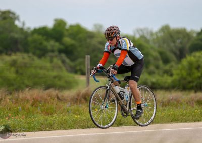 Velo cyclist riding on scenic countryside road with lush greenery, promoting Bluebonnet Express bike services and outdoor adventure.