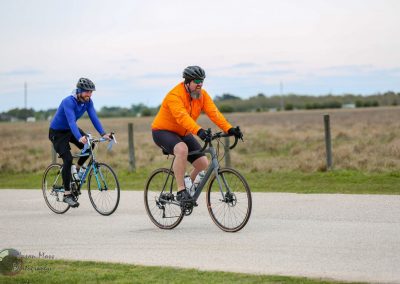 Brightly colored cyclists riding on a scenic outdoor trail with open fields and a cloudy sky in the background.