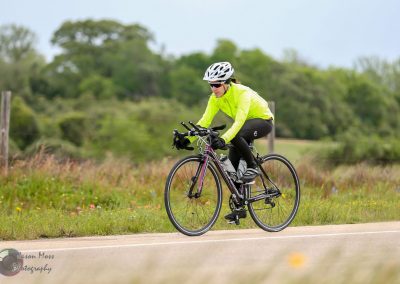 Bright yellow cycling jacket on a woman riding a road bike through open countryside for Bluebonnet Express.