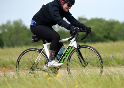 Vibrant woman riding a Cervelo road bike on a scenic trail, promoting Bluebonnet Express cycling events and outdoor adventure.