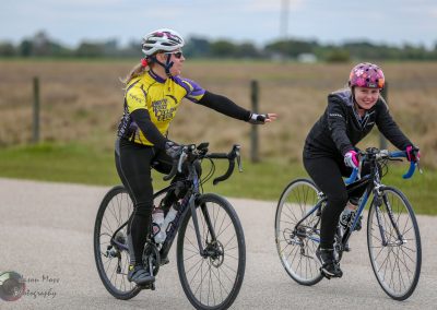 Adult woman cycling with a young girl, both wearing helmets, smiling and enjoying riding bikes outdoors.
