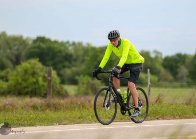 Bright yellow cycling jacket on male cyclist riding a black e-bike on rural road, outdoor biking adventure, Bluebonnet Express.