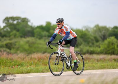 High-performance cyclist riding on a rural road in a Bluebonnet Express cycling jersey and helmet.