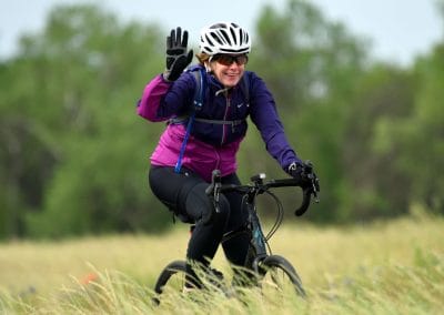 Bright woman cycling outdoors on a mountain bike, wearing helmet and gear, with a smiling face, in a lush green field.