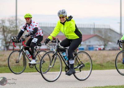 Bright yellow cycling jacket on female cyclist riding Bluebonnet Express bike, smiling and waving.