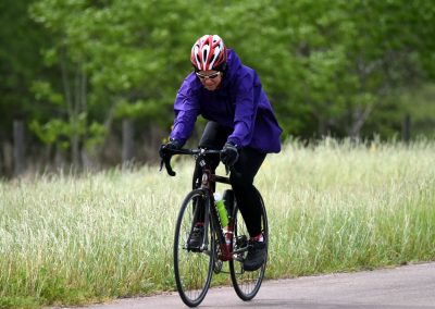 Bright woman riding a bike on a trail in nature, wearing a purple jacket, helmet, and sunglasses.