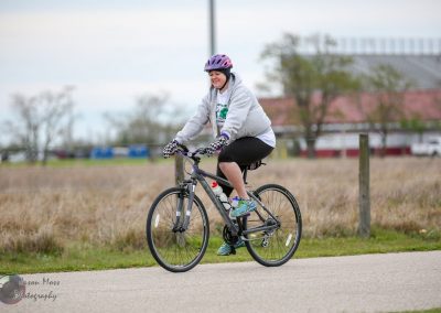 Smiling woman riding a bicycle outdoors during daytime, promoting Bluebonnet Express cycling event.