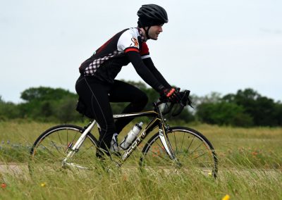 Vibrant cyclist riding mountain bike through grassy field with trees in background, promoting Bluebonnet Express cycling adventures.