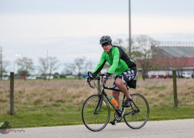 Man riding a bicycle on a paved trail, wearing a green jacket and helmet, for Bluebonnet Express cycling event.