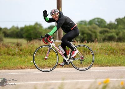 Vastly experienced cyclist waving while riding a high-performance road bike on scenic rural road.