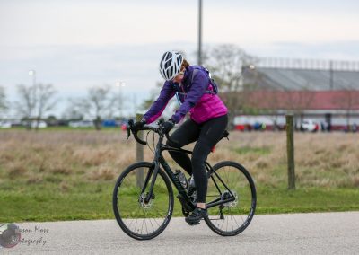 Bright female cyclist riding a mountain bike outdoors with a stadium in the background, promoting Bluebonnet Express biking events.