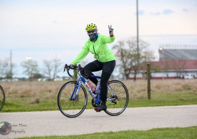 Bright green cycling jacket with Bluebonnet Express logo, cyclist waving, on scenic bike trail.