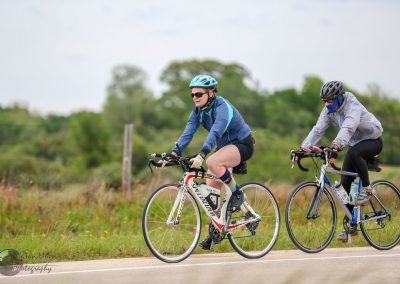 Bright blue and gray cycling jackets, helmets, and sporty gear on two cyclists riding on open road in green landscape.
