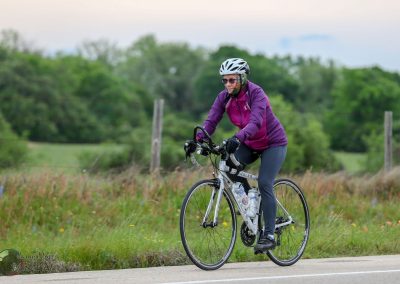 Vibrant woman cycling outdoors on a scenic trail with lush greenery and a clear sky, promoting Bluebonnet Express bike services.