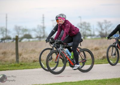 Bright female cyclist riding on a trail with outdoor scenery in the background, wearing a helmet and athletic gear.