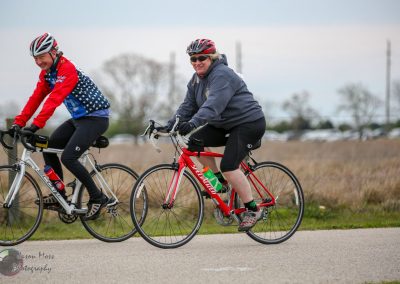 Joyful cyclists riding the Bluebonnet Express bike route with scenic open fields and cloudy skies in the background.