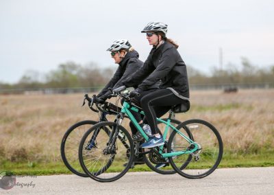 Women riding electric bikes outdoors on a paved trail in a rural area.