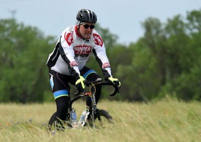 Rider in BMX biking gear cycling on a grassy trail with green trees in background.