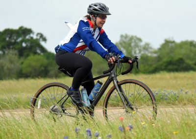 Bold woman cycling on trail with a carbon bike in lush green field, sporting Bluebonnet Express cycling gear.