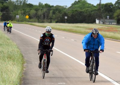 Brightly dressed cyclists riding on a scenic highway during daytime, promoting Bluebonnet Express bike tour.