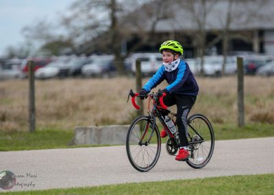 Child riding a bicycle during daytime, wearing a neon green helmet, blue jacket, and red shoes.