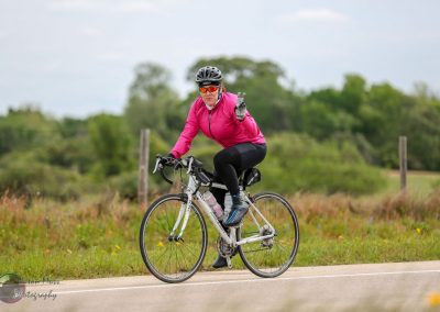 Vegan cyclist in pink jacket making peace sign while riding on country road during daytime.
