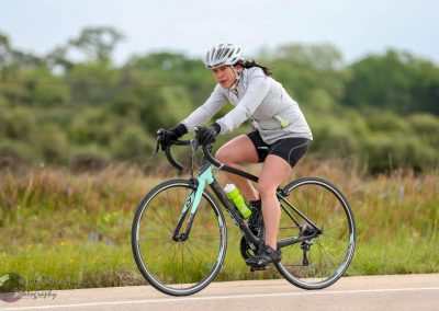 Woman riding a road bike with helmet and athletic gear through a scenic outdoor landscape.