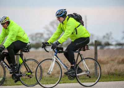 Bright yellow cycling jackets on two riders enjoying a ride on a rural road with a cloudy sky.