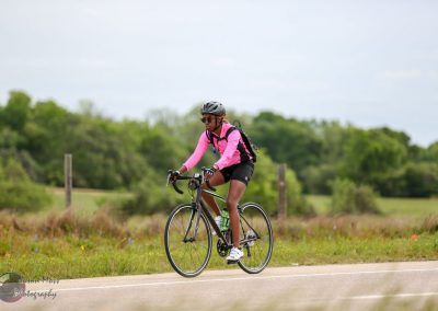 Bright female cyclist riding on rural road in pink athletic gear and helmet for Bluebonnet Express cycling event.