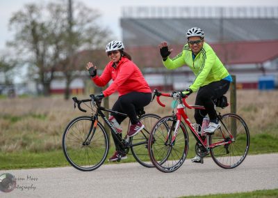 Brightly dressed cyclists riding bikes outdoors, smiling and waving, promoting fitness and active transportation.