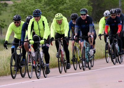 Highway cycling group wearing yellow and dark gear, riding together on scenic road, promoting Bluebonnet Express cycling event.