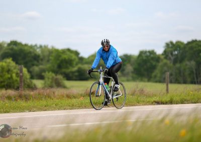 Mountain bike rider on rural road in Texas countryside under blue sky, promoting Bluebonnet Express cycling tour.