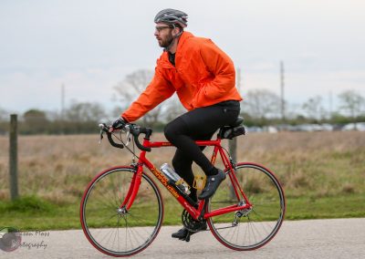 Bright red road bike with a cyclist in orange jacket riding outdoors in cloudy weather.