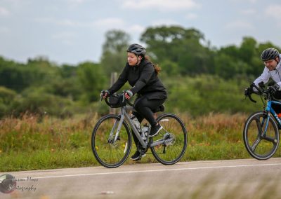 Female cyclist riding an e-bike along a scenic rural trail with lush greenery and colorful wildflowers.