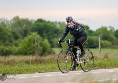 Woman smiling riding a bicycle outdoors in a scenic countryside setting, promoting Bluebonnet Express cycling tours.