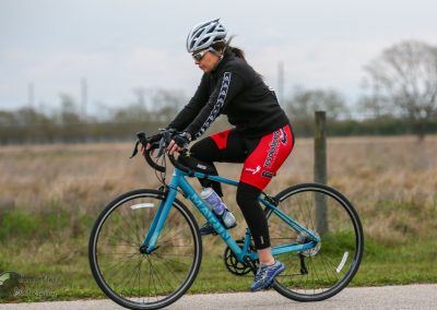 Woman riding blue road bike with helmet and cycling gear on rural trail during daytime.