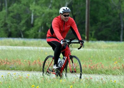 Brightly dressed cyclist riding on a scenic trail surrounded by green trees and wildflowers, promoting outdoor biking with Bluebonnet Express.