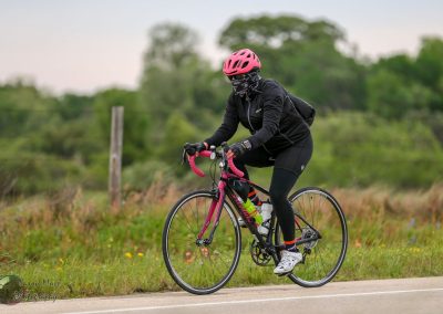 Vegan cyclist riding on a rural road with a pink bike, wearing protective gear and black athletic clothing.