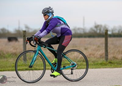 Womens cycling trail on country road with mountain bike, wearing purple jacket, black leggings, and helmet.